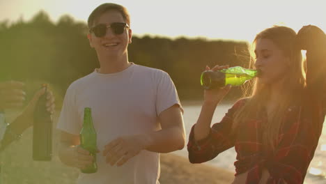 Loving-couple-celebrate-the-end-of-the-semester-with-beer-and-pop-music-on-the-beach-with-their-friends.-Girl-is-dancing-on-the-open-air-party-and-drinking-beer-at-sunset-in-hot-summer-evening.